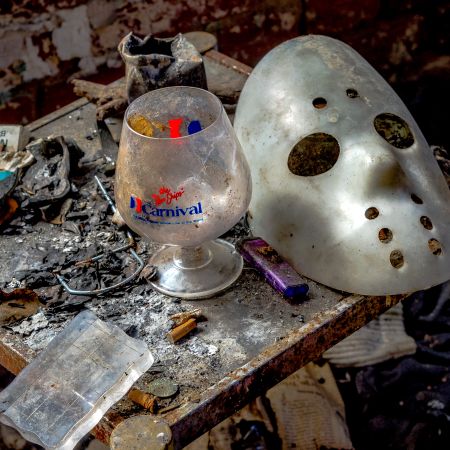 Abandoned artifacts including white hockey mask and carnival cruise glass on table in derelict greenhouse complex.