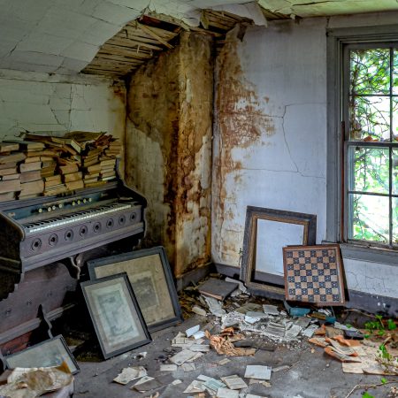 Abandoned attic in rural home with old organ, stacks of old books and prints and crumbling ceiling and green vines.