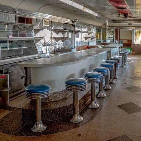 Abandoned 1950s style diner with chrome stools and backsplash, diamond patterned floor and row of booths.