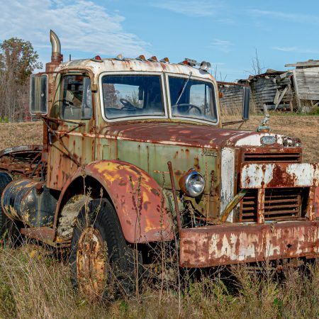 Vintage Semi Mack truck in a field with abandoned building in rear.