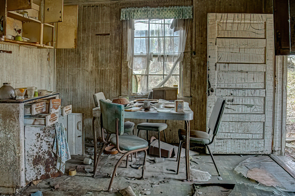 Derelict farmhouse kitchen with tables and chairs, peeling paint, open door and dishtowels on the sink.