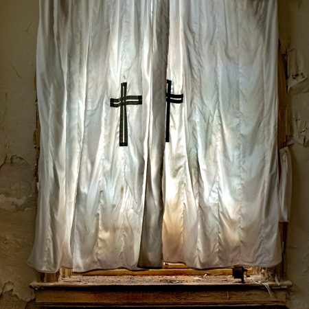 A curtain with embroidered crosses illuminates afternoon sunlight within an abandoned prison chapel.